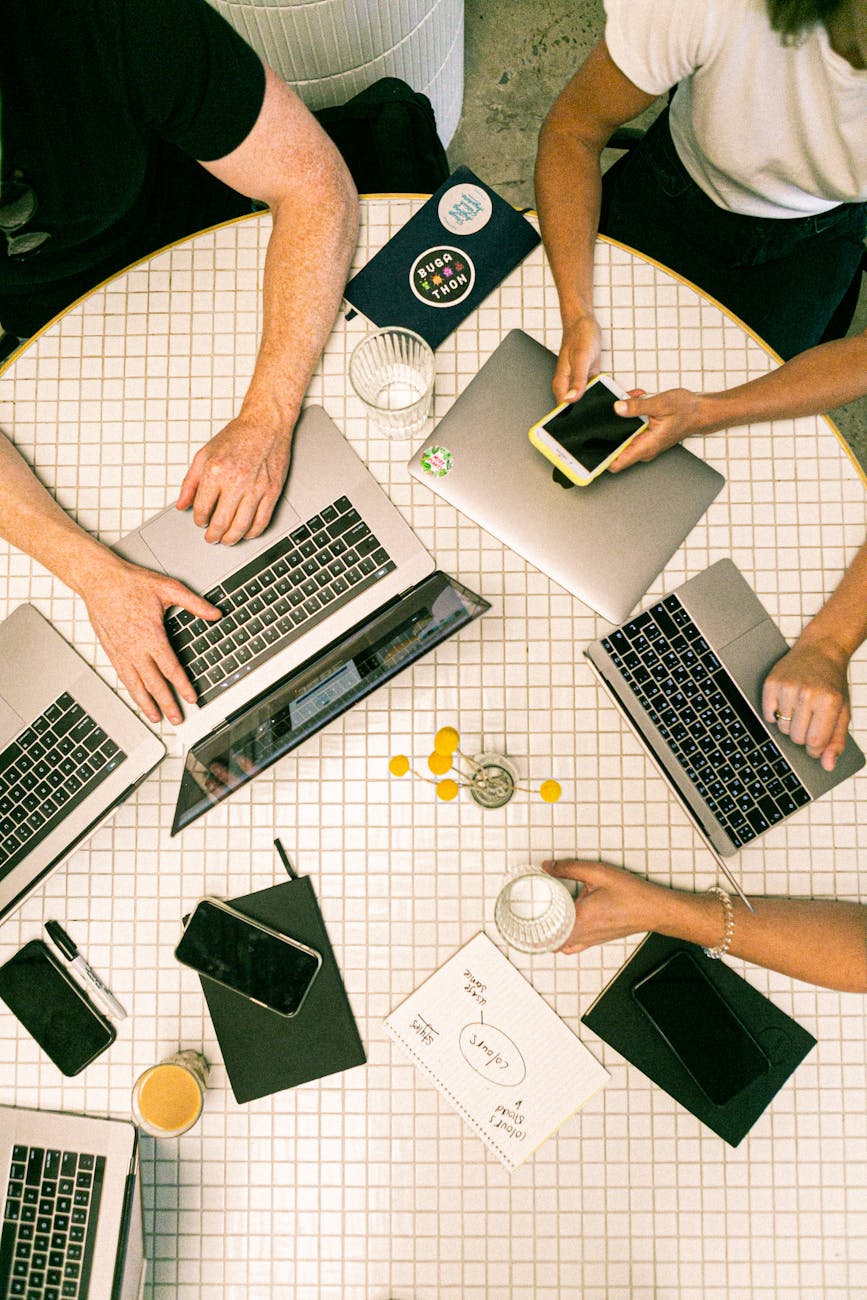 Overhead view of a group working around a round table, using laptops and a smartphone, with drinks, notebooks, and a small vase of yellow flowers scattered across the tiled surface.