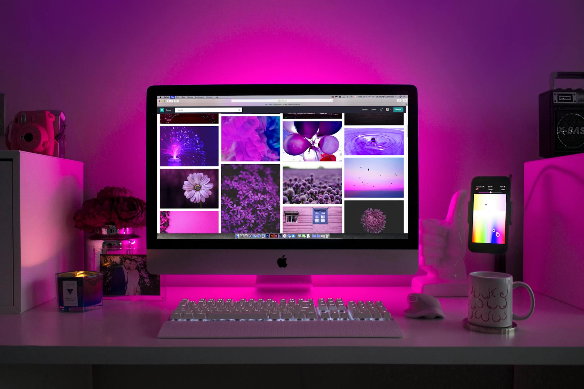 Desktop computer displaying a gallery of vibrant purple and pink images, set on a desk with glowing pink backlighting, keyboard, coffee mug, and smartphone.