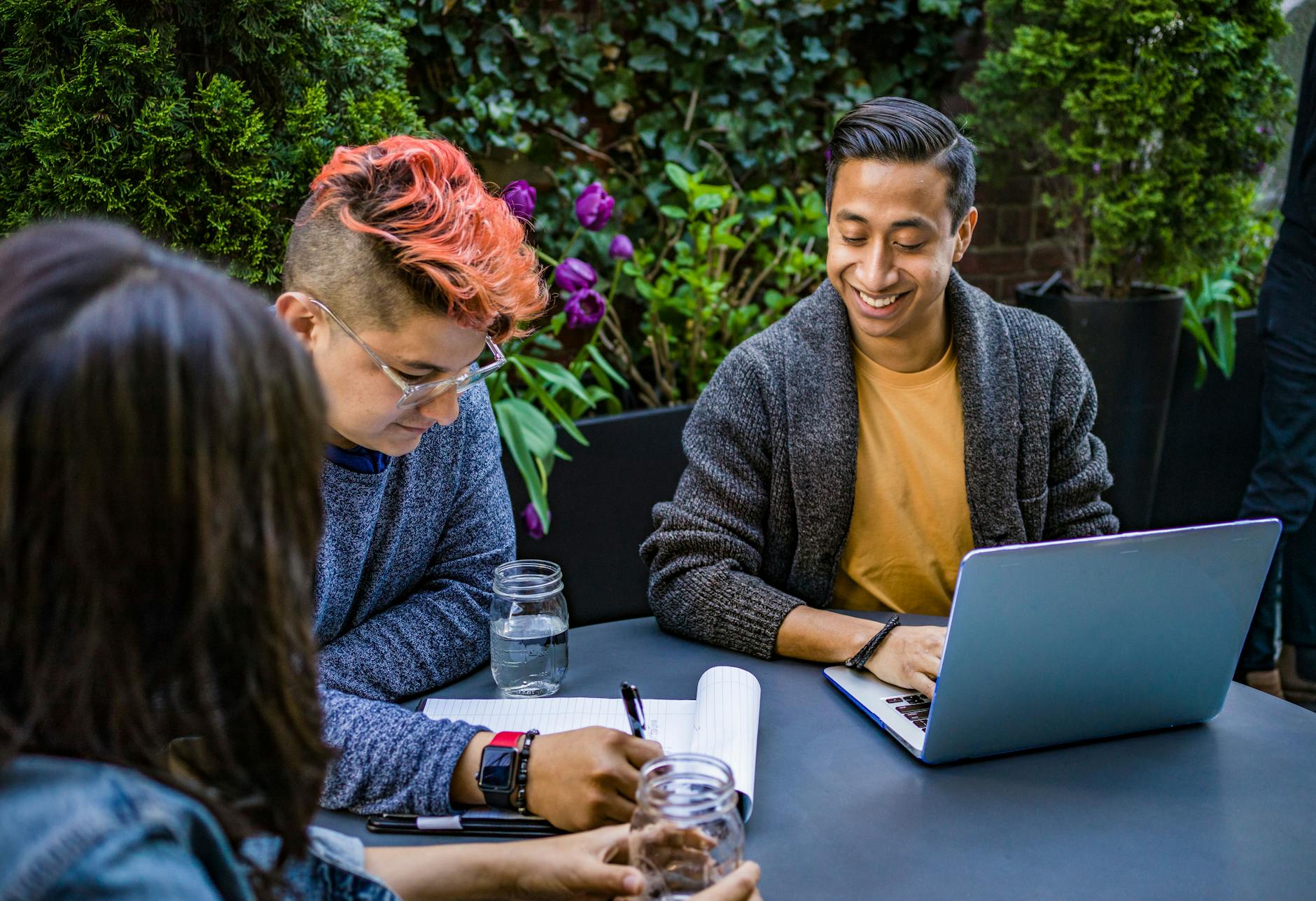 Three colleagues collaborate outdoors at a table surrounded by greenery. One person types on a laptop while another takes notes, creating a relaxed and friendly atmosphere that reflects teamwork, creativity, and open communication.