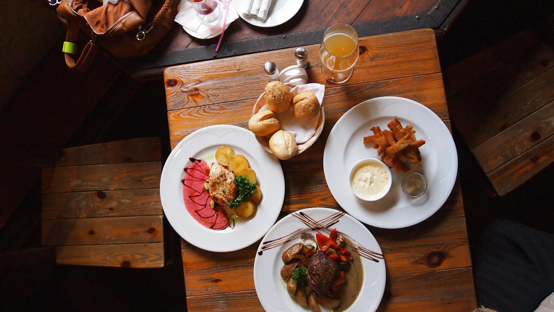 Overhead view of a rustic restaurant table set with three plated entrées (sliced roast with potatoes, seared chicken, and steak with vegetables), a basket of rolls, fries with dipping sauce, and a drink.