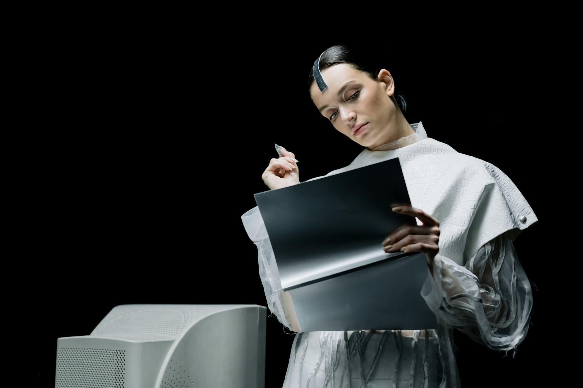 woman holding a pen while looking at a folder Futuristic writer reviewing notes beside a retro computer.