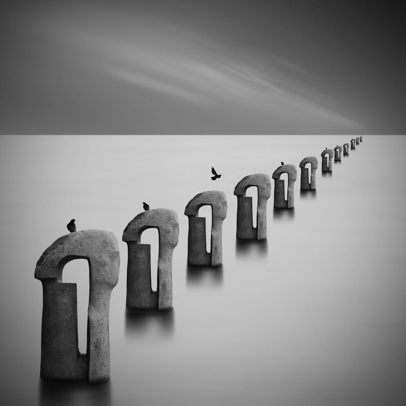 A minimalist black-and-white photo shows a calm, glassy sea. A repeating line of weathered stone arch-shapes stretches from the foreground to a vanishing point on the right. Small birds perch on several stones; one bird is caught mid-flight above the smooth water. The scene feels quiet and surreal.