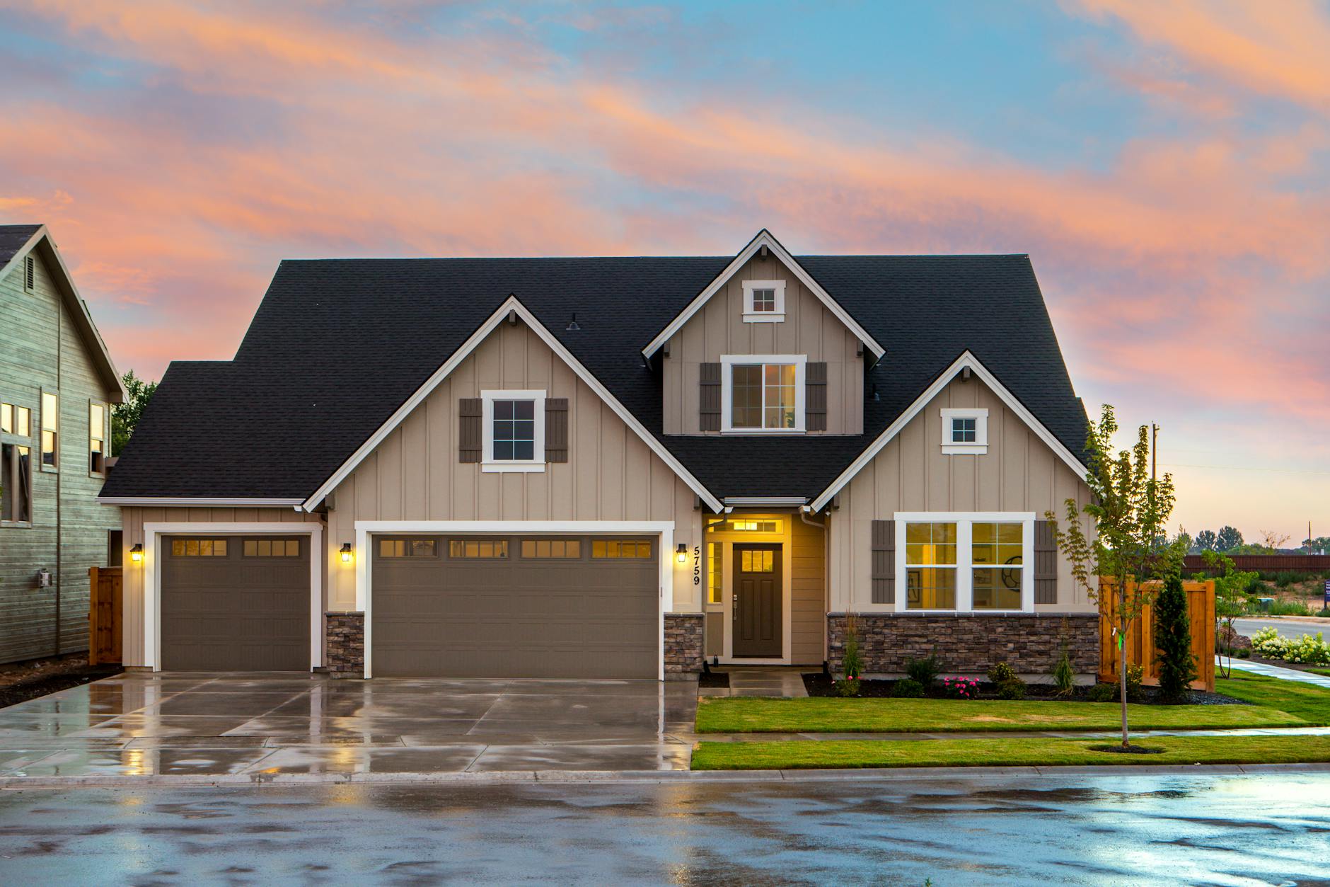A modern suburban house with a three-car garage, gray siding, white trim, and a dark shingled roof. The front door is warmly lit and framed by stone accents along the lower portion of the exterior. Large windows on both floors glow with interior light, and a neatly manicured lawn with young trees and shrubs lines the front yard. The driveway is wet, reflecting the house and the soft colors of a pink and blue sunset sky.