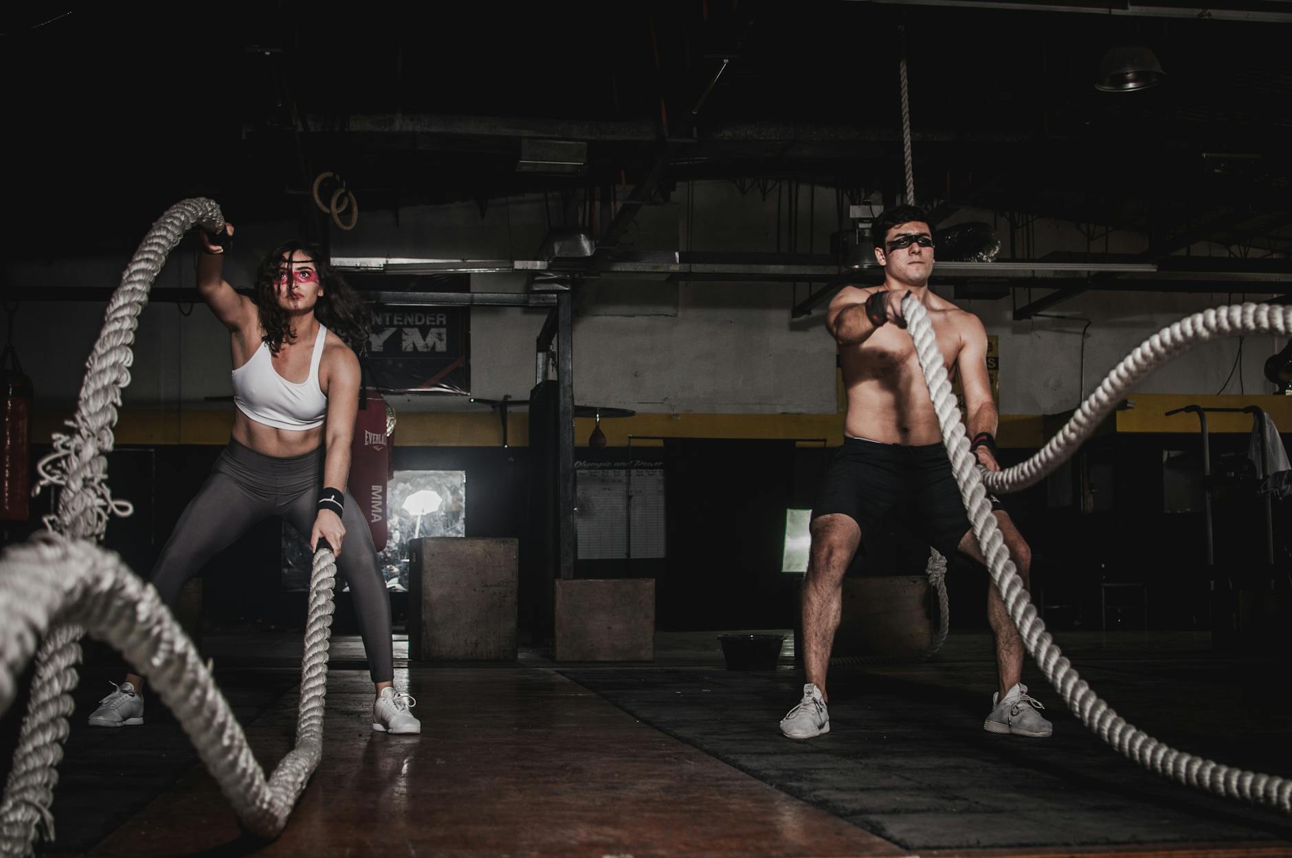 Inside a dim, industrial-style gym, a woman and a man perform synchronized battle rope waves—knees bent, cores engaged—surrounded by boxes, weights, and worn concrete floors.