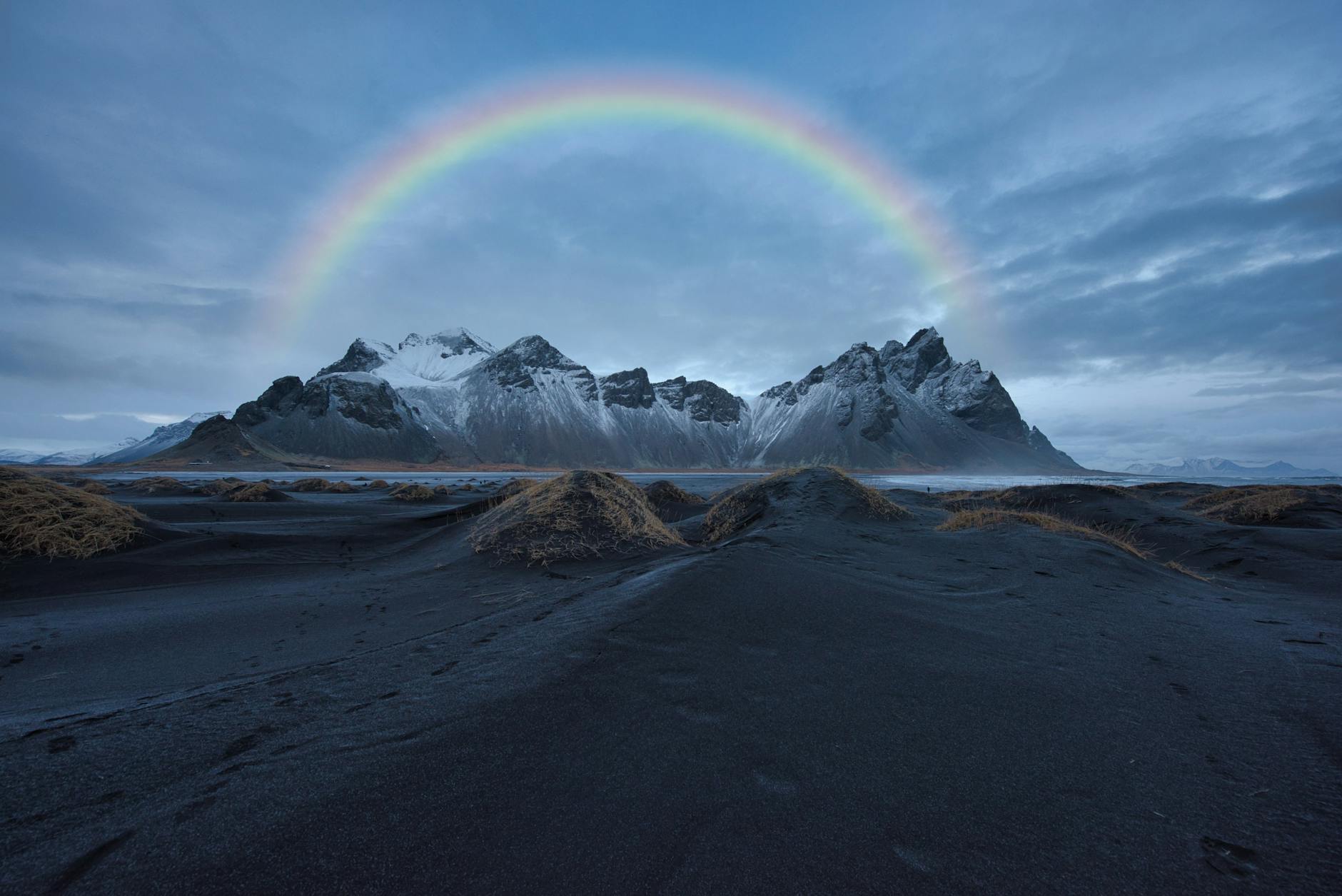 A bright rainbow arcs over snow-covered mountains and black sand dunes under a cloudy blue sky.