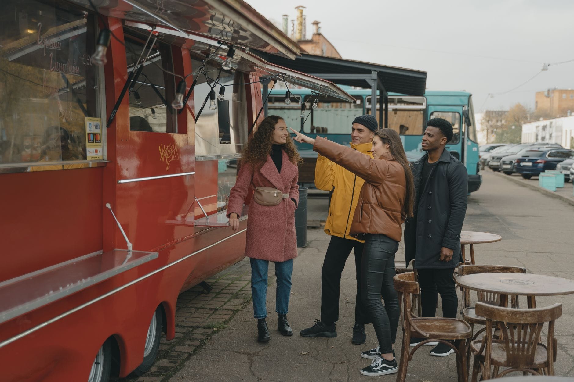 Four friends smile and take a photo while ordering food from a red food truck at an outdoor dining area.